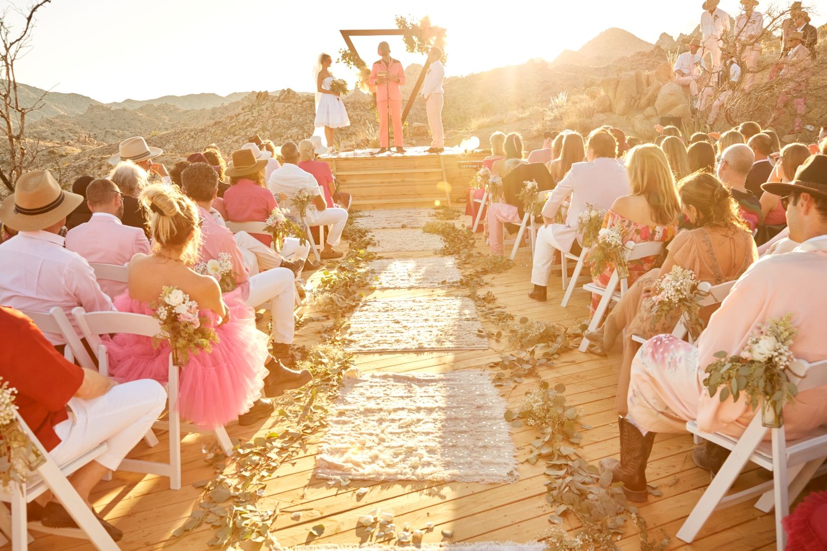 A wedding ceremony at The Triangle at ESCAPE — aisle through eucalyptus and roses to the wood arch, golden hour, Pioneertown mountains behind